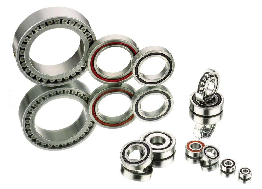 A variety of metal ball bearings of different sizes and types are arranged on a reflective surface. The bearings showcase intricate designs, including cylindrical roller and ball bearings, with some featuring red seals.